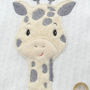 Könnte beinhalten: Nahaufnahme einer gestickten Giraffen-Applikation in Beige und Grau. Die Giraffe hat ein freundliches Gesicht mit schwarzen Augen und einem roten Lächeln. Die Flecken der Giraffe sind grau und der Hintergrund ist weiß.