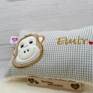 May include: A rectangular pillow with a gray and white checkered pattern. The pillow features an embroidered monkey face on one side and the name "Emir" in gold thread. A small red heart is next to the name. The pillow is resting on a white surface.