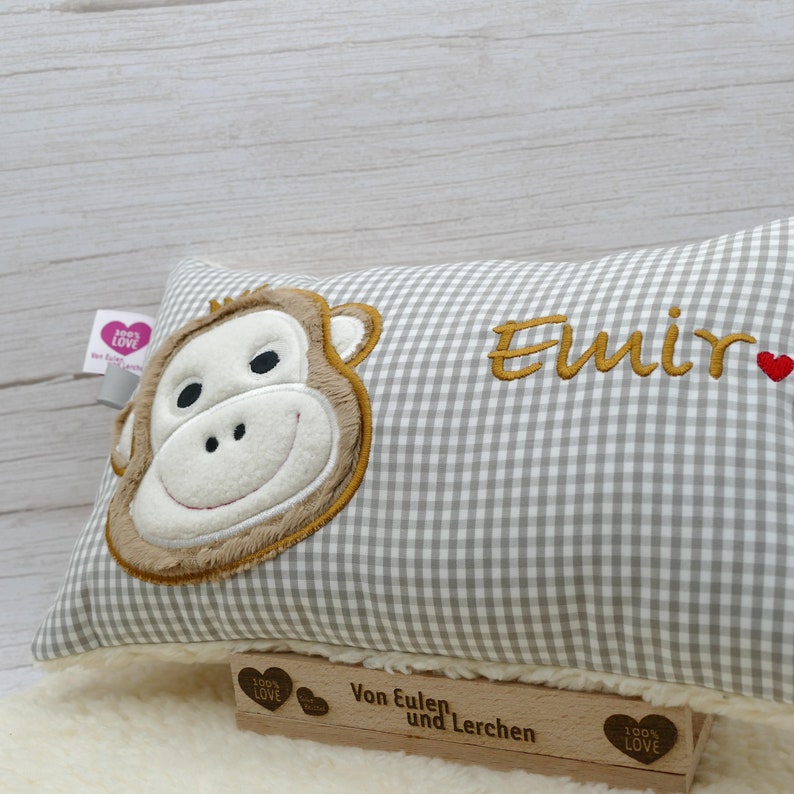 May include: A rectangular pillow with a gray and white checkered pattern. The pillow features an embroidered monkey face on one side and the name "Emir" in gold thread. A small red heart is next to the name. The pillow is resting on a white surface.