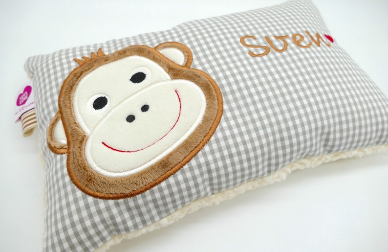 May include: A rectangular pillow with a gray and white gingham pattern. The pillow features a large, embroidered monkey face in shades of brown, cream, and black. The name "Sven" is embroidered in brown. The pillow has a soft, cream-colored border.