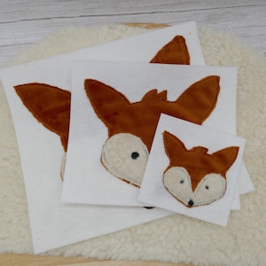Könnte beinhalten: Drei Filz-Applikationen mit Fuchsgesichtern in verschiedenen Größen. Die Applikationen sind braun mit weißem Fell und schwarzen Augen. Die Applikationen befinden sich auf einem weißen Hintergrund.