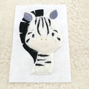 Könnte beinhalten: Ein weiß-schwarzes Zebra-Applikationsmotiv mit schwarzer Mähne und grauen Ohren. Das Zebra ist auf einem weißen Filzgrund gestickt.