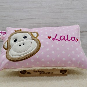 May include: Pink rectangular pillow with a monkey face design and the name "Lala" embroidered in pink. The pillow has a white star pattern and a brown wooden sign that says "Von Eulen und Lerchen".