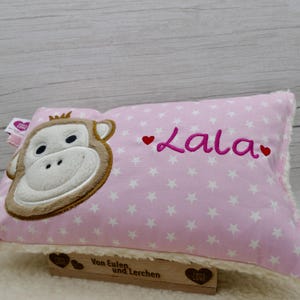May include: Pink pillow with white star pattern and a monkey face applique. The name "Lala" is embroidered in pink with red hearts. The pillow has a fluffy white border and a wooden base.