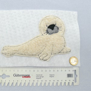 Könnte beinhalten: Ein weißer Plüsch-Seehunds-Applikationen mit schwarzen Augen und einer grauen Nase. Der Seehund liegt auf einer weißen Oberfläche mit einem Lineal und einer Münze.