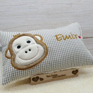 May include: A rectangular pillow with a gray and white gingham pattern. The pillow features an embroidered monkey face in shades of brown, tan, and white. The name "Elmir" is embroidered in gold thread, with a small red heart. A wooden block with text is at the bottom.