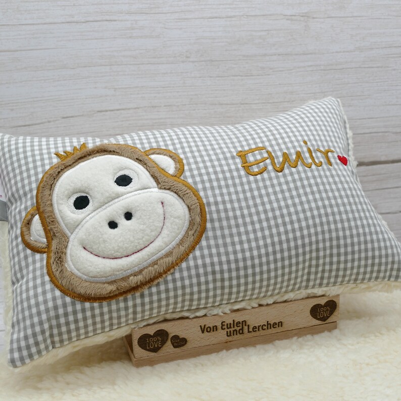 May include: A rectangular pillow with a gray and white gingham pattern. The pillow features an embroidered monkey face in shades of brown, tan, and white. The name "Elmir" is embroidered in gold thread, with a small red heart. A wooden block with text is at the bottom.