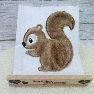 Könnte beinhalten: Ein weißes Filzquadrat mit einer braunen Plüsch-Eichhörnchen-Applikation, weißem Gesichtsfleck und schwarzem Auge. Der Filz steckt in einem hellen Holzständer, der mit "Von Eulen und Lerchen" und zwei Herzen mit "100% LOVE" graviert ist.