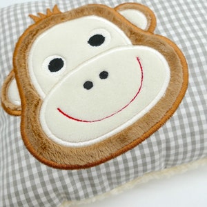 May include: A gray and white gingham patterned pillow featuring a plush monkey face with brown fur, a cream-colored face, and a red stitched smile. The pillow has a soft, fluffy border.