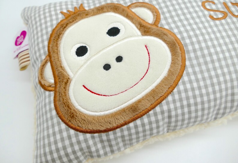 May include: A gray and white gingham patterned pillow featuring a plush monkey face with brown fur, a cream-colored face, and a red stitched smile. The pillow has a soft, fluffy border.
