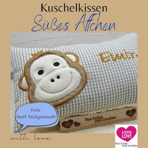 Könnte beinhalten: Ein grau-weiß kariertes Kissen mit einem braunen Affengesicht, das darauf gestickt ist. Das Kissen trägt den Namen "Emir", der in goldener Farbe gestickt ist. Das Kissen liegt auf einer Holzoberfläche mit einem herzförmigen Etikett, auf dem "100% LOVE" und der Text "Von Eulen und Lerchen" steht.