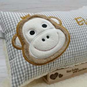May include: A rectangular pillow with a gray and white checkered pattern. The pillow features an embroidered monkey face with brown, white, and black details. The pillow is resting on a white, fluffy surface.