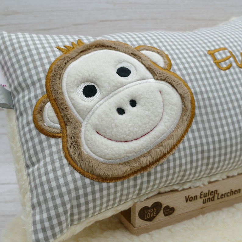 May include: A rectangular pillow with a gray and white checkered pattern. The pillow features an embroidered monkey face with brown, white, and black details. The pillow is resting on a white, fluffy surface.