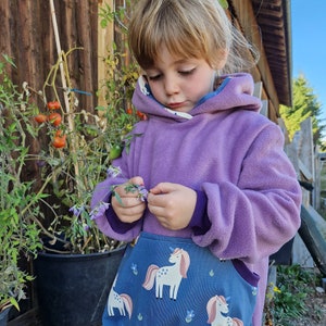 May include: A purple fleece hoodie with a blue unicorn pocket. The pocket features white unicorns with pink manes and tails, and blue flowers on a blue background. The hoodie has a blue lining in the hood and cuffs.