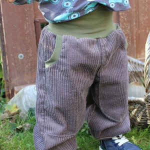 May include: A toddler wearing a pair of grey corduroy trousers with a green waistband and a blue and white shirt with a tractor print. The toddler is wearing blue trainers with white laces.