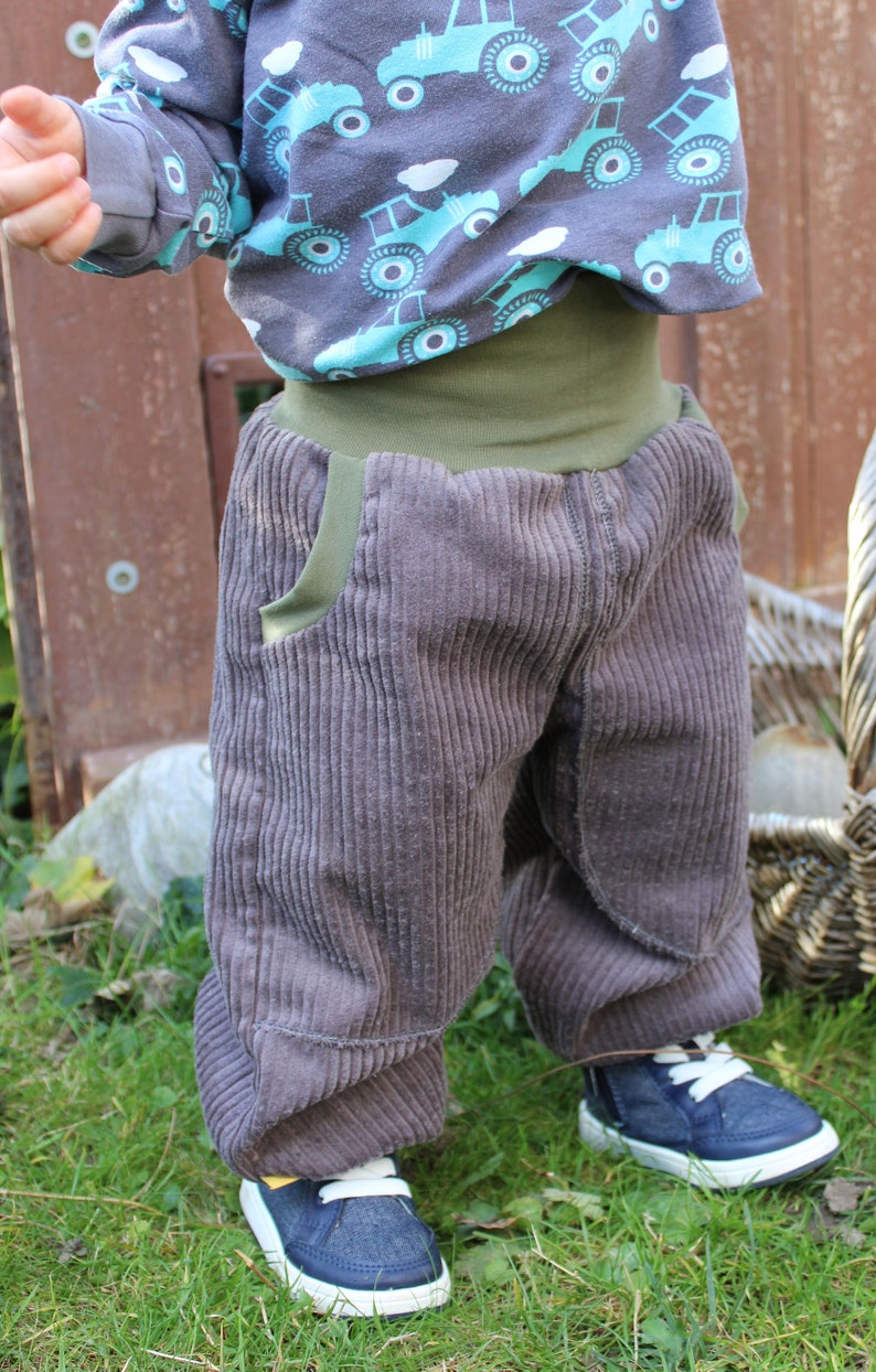 May include: A toddler wearing a pair of grey corduroy trousers with a green waistband and a blue and white shirt with a tractor print. The toddler is wearing blue trainers with white laces.