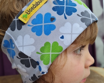 May include: A gray headband with a white, blue, and green four-leaf clover pattern. The headband has a yellow label that says "Boedabutik".