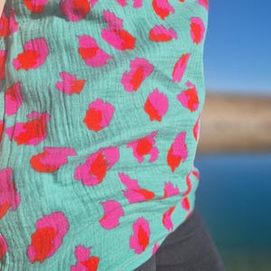 Könnte beinhalten: Ein türkisfarbenes Oberteil mit einem auffälligen Muster aus rosa und roten abstrakten Formen. Der Stoff hat ein strukturiertes Aussehen. Das Oberteil wird mit einer dunkelgrauen Hose kombiniert, was einen auffälligen Kontrast erzeugt. Der Hintergrund zeigt einen klaren blauen Himmel.