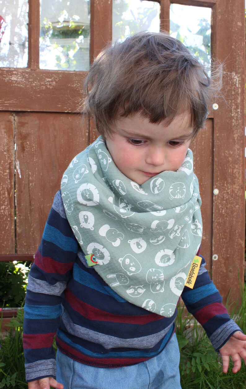 Puede incluir: Un ni&ntilde;o peque&ntilde;o con una camisa de manga larga a rayas azules y rojas con un pa&ntilde;uelo verde con caras de animales blancas y el texto "Boeddhabutik".