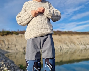 May include: A child wearing a navy blue beanie, a beige knit sweater, and gray pants with blue patterned accents. The child is standing on a rocky surface with a body of water in the background.