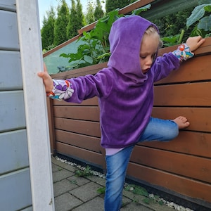 May include: A young person wearing a purple hooded sweatshirt with colorful floral print on the sleeves and blue jeans. The person is standing with one leg raised and is leaning against a wooden fence.