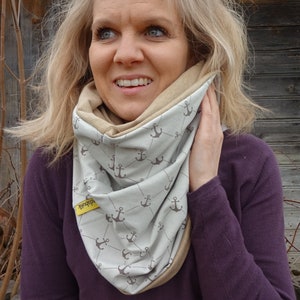 May include: A woman wearing a light purple long-sleeved shirt with a beige and light blue infinity scarf featuring a repeating anchor pattern. The scarf has a yellow tag that says "Seaduck".