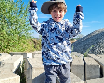 Könnte beinhalten: Ein kleiner Junge trägt einen blau-weißen Kapuzenpullover mit Dinosaurier-Motiv, eine graue Baseballkappe und eine blaue Jeans. Er steht auf einer Betonfläche mit erhobenen Armen.