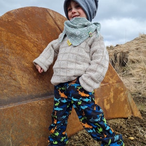 May include: A young child wearing a light blue beanie, a light green scarf, a beige sweater, and blue pants with a shark print. The child is standing next to a large rusty metal object.