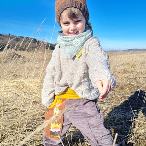 May include: A young child wearing a brown knitted hat with orange stripes, a light blue bandana, a cream sweater, and grey corduroy pants with a yellow waistband and a colorful cartoon print on the pocket. The child is standing in a field of tall dry grass.