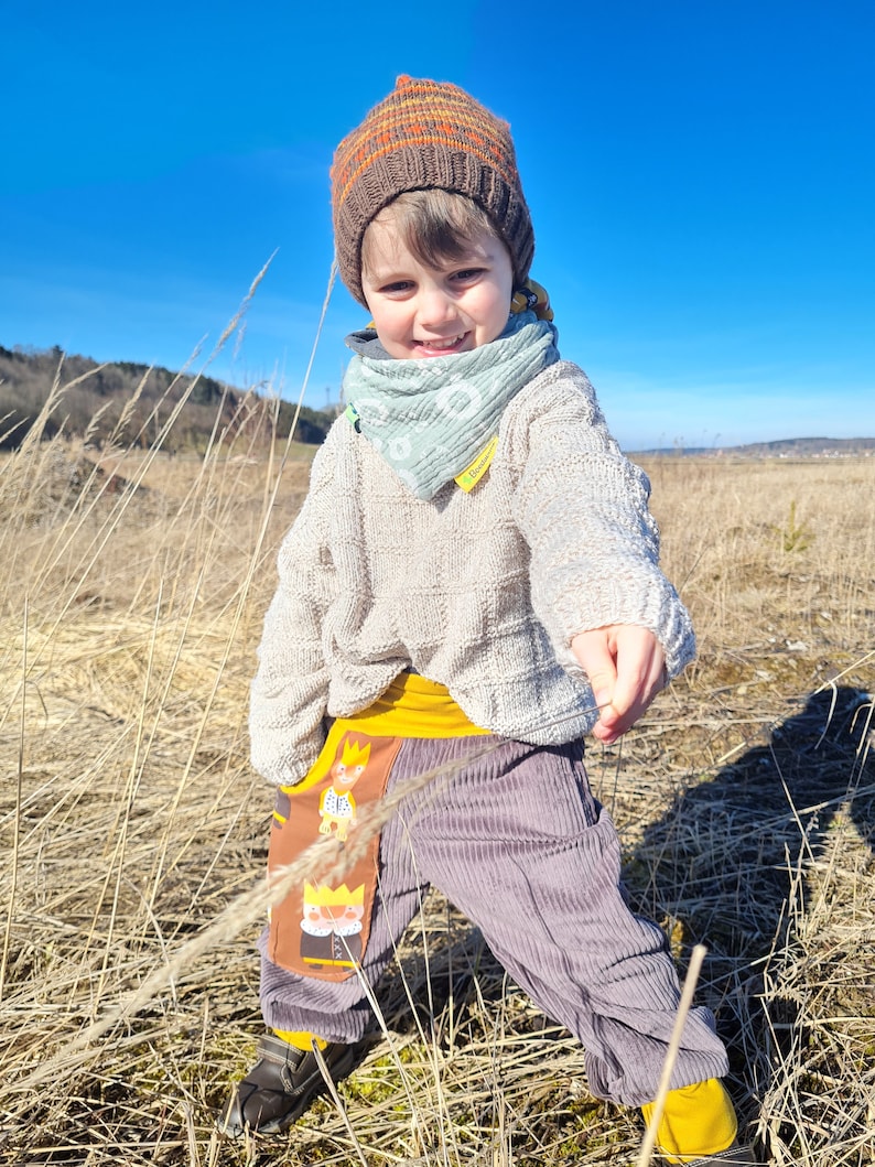 May include: A young child wearing a brown knitted hat with orange stripes, a light blue bandana, a cream sweater, and grey corduroy pants with a yellow waistband and a colorful cartoon print on the pocket. The child is standing in a field of tall dry grass.