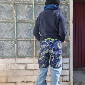 Op de afbeelding: Een kind draagt een blauwe trui en een blauwe spijkerbroek met een ruimtethema in groen en blauw. Het kind draagt een zwarte beanie met een wit logo. Het kind staat voor een bakstenen muur met een glas-in-loodraam.