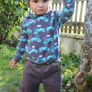 May include: A young child wearing a grey and brown knitted hat, a grey long-sleeved shirt with a blue and white tractor print, grey corduroy trousers, and black trainers. The child is standing in a grassy area with a wooden fence and a tree in the background.