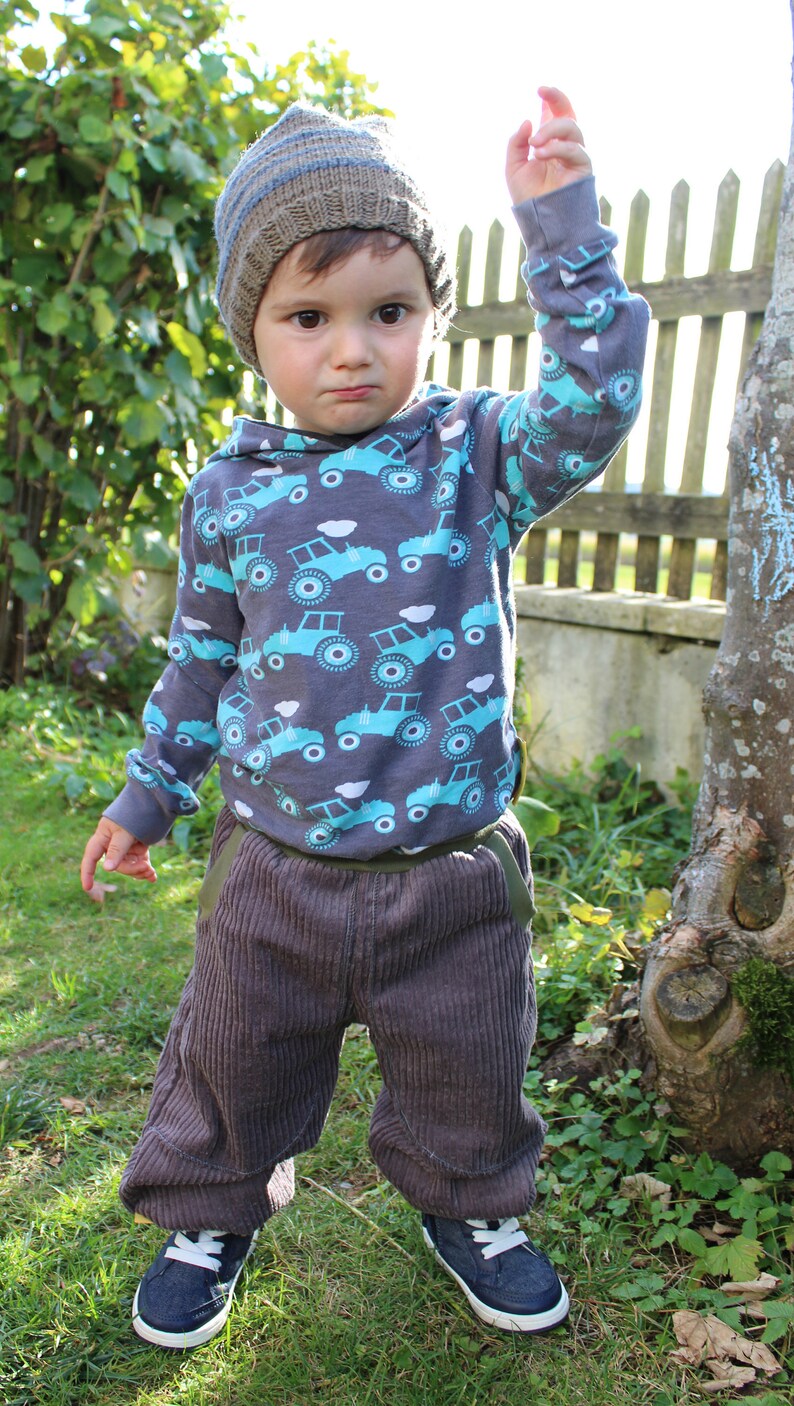 May include: A young child wearing a grey and brown knitted hat, a grey long-sleeved shirt with a blue and white tractor print, grey corduroy trousers, and black trainers. The child is standing in a grassy area with a wooden fence and a tree in the background.