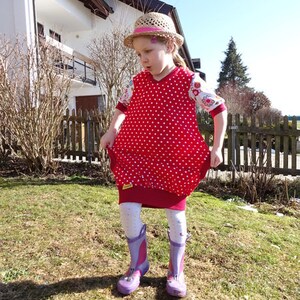 Puede incluir: Una ni&ntilde;a peque&ntilde;a lleva un vestido de lunares rojos y blancos con estampado floral blanco en las mangas. Lleva un sombrero de paja con una cinta rosa y botas de lluvia moradas.