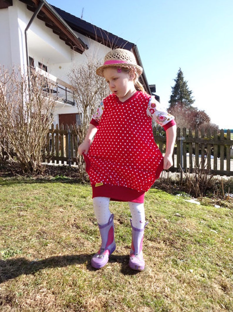 Puede incluir: Una ni&ntilde;a peque&ntilde;a lleva un vestido de lunares rojos y blancos con estampado floral blanco en las mangas. Lleva un sombrero de paja con una cinta rosa y botas de lluvia moradas.