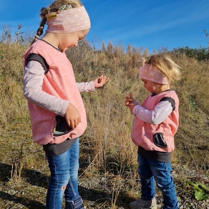 May include: Two children wearing pink and grey vests with matching pink headbands with white stars. The children are standing in a grassy field with a blue sky in the background.
