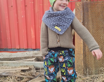 May include: A young child wearing a green beanie, a gray and white bandana, a brown sweater, and black pants with a dinosaur print. The child is standing in front of a red metal wall.
