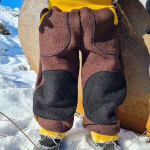 Può includere: Pantaloni in pile marrone con ginocchiere nere e bordino giallo. I pantaloni sono indossati da un bambino in piedi sulla neve.