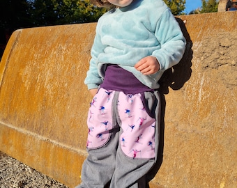 May include: A young child wearing a light blue sweatshirt, a purple waistband, and gray pants with pink and white ski-themed pockets. The child is standing next to a large, rusty metal object.