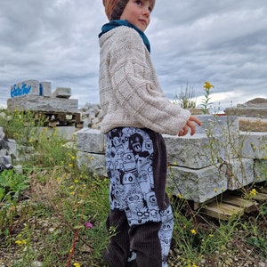 Op de afbeelding: Een kind draagt ​​een bruine en oranje gebreide muts, een witte gebreide trui en een bruine corduroy broek met een zwart-witte cartoonprint. Het kind staat in een veld met rotsen en groene planten.
