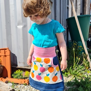 Könnte beinhalten: Ein kleines Kind trägt ein türkisfarbenes T-Shirt und einen Rock mit einem rosa Bund und einem weißen Hintergrund mit orangefarbenen und rosafarbenen Fruchtmotiven. Der Rock hat einen blauen Jeans-Saum.