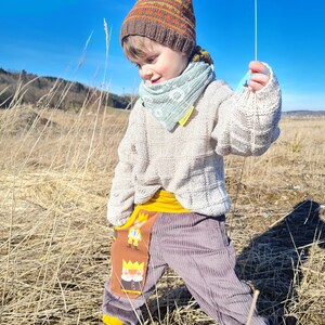 May include: A young child wearing a brown and orange knit hat, a light blue bandana, a white sweater, and gray corduroy pants with a yellow waistband and a colorful cartoon character patch. The child is standing in a field of tall dry grass.