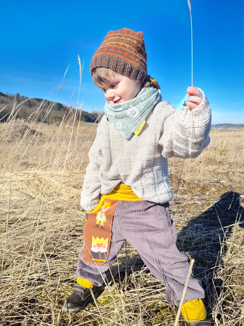 May include: A young child wearing a brown and orange knit hat, a light blue bandana, a white sweater, and gray corduroy pants with a yellow waistband and a colorful cartoon character patch. The child is standing in a field of tall dry grass.