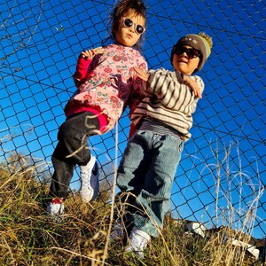 May include: Two children wearing sunglasses and hats stand in front of a chain link fence. The girl is wearing a pink and white floral shirt and black pants. The boy is wearing a striped sweater and blue jeans.