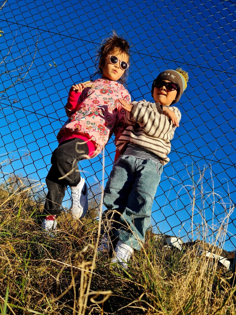 May include: Two children wearing sunglasses and hats stand in front of a chain link fence. The girl is wearing a pink and white floral shirt and black pants. The boy is wearing a striped sweater and blue jeans.