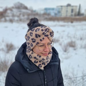 May include: A tan and black leopard print headband and neck warmer set. The soft, fuzzy fabric is a neutral color with dark brown spots. The person is wearing a black jacket and glasses in a snowy outdoor setting.