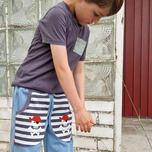 May include: A child wearing a gray t-shirt with a pocket and blue denim pirate-themed pants. The pants have striped pockets with a pirate skull and crossbones design. The child is standing outside.