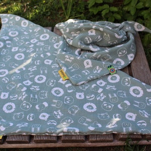 Puede incluir: Una manta de muselina verde con estampado de animales blancos. La manta presenta una variedad de animales, incluidos leones, osos, mapaches y conejos. La manta est&aacute; doblada por la mitad y tiene una etiqueta amarilla con la palabra "Swaddling" en ella.