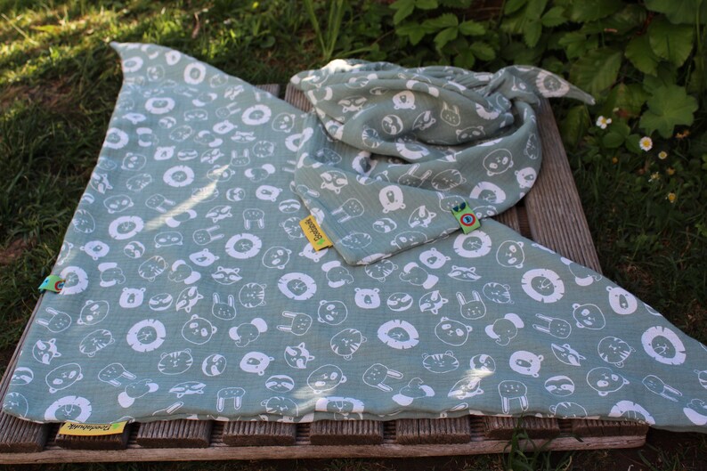 Puede incluir: Una manta de muselina verde con estampado de animales blancos. La manta presenta una variedad de animales, incluidos leones, osos, mapaches y conejos. La manta est&aacute; doblada por la mitad y tiene una etiqueta amarilla con la palabra "Swaddling" en ella.