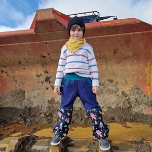 May include: A young person wearing a white and blue striped jumper, a yellow scarf, and a black beanie stands in front of a large red construction vehicle. They are wearing blue jeans with a rocket ship pattern on the legs.